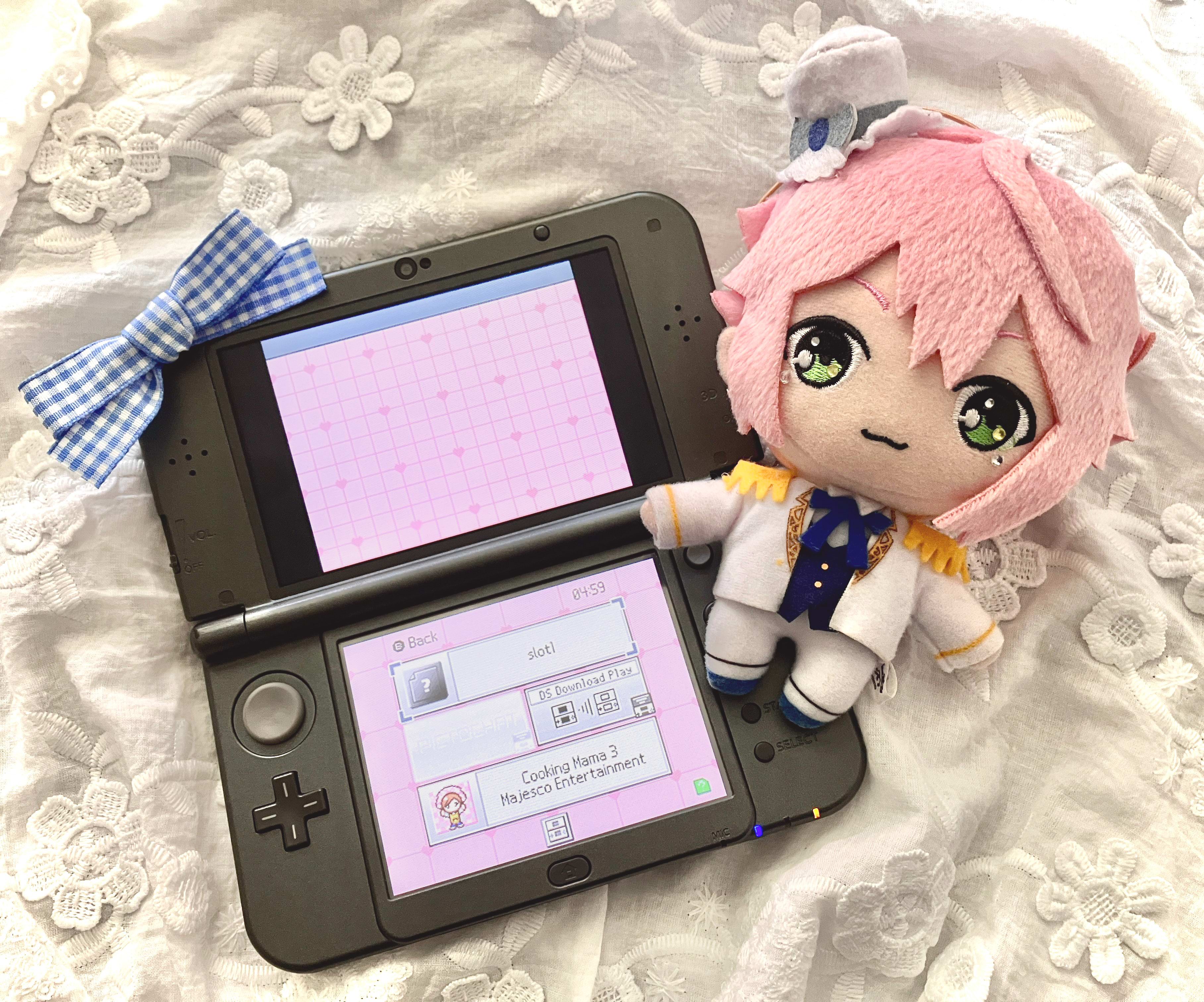 Tori Himemiya plush lying right to a 3ds displaying a custom pink dsi theme, to the left is a blue gingham ribbon. Underneath is a white cloth embroidered with flowers.